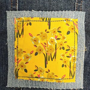 May include: Close-up of a denim jacket with a yellow floral patch. The patch is a square of yellow fabric with a pattern of brown and pink flowers. The patch is sewn onto the jacket with a denim border.