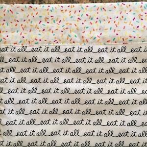 May include: A white pillowcase with a top section covered in colorful sprinkles. The lower section is white with the words "eat it all" repeated in a black, cursive font. The pillowcase is likely for a bed or couch.