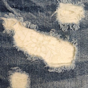 May include: Blue denim fabric featuring distressed areas with frayed edges. Beneath the large central tear and smaller holes, a cream-colored fabric patch is visible, decorated with a delicate, light-colored floral and geometric line pattern.