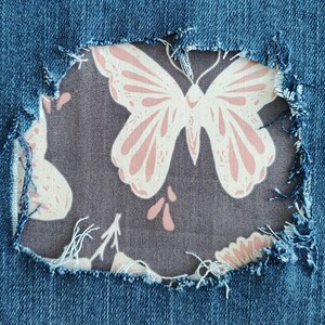 May include: A blue denim fabric with a large, distressed, frayed hole revealing a dark gray fabric patch underneath. The patch features a repeating pattern of light pink butterflies and small floral elements printed on the dark gray background.