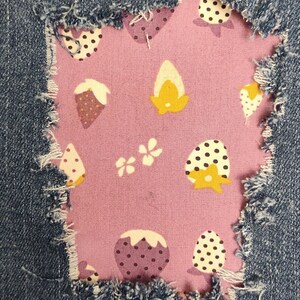 May include: A pair of blue denim jeans featuring a rectangular distressed patch area. The opening reveals a light purple fabric lining decorated with a repeating pattern of white and yellow strawberries, black polka dots, and small white flowers.
