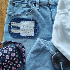 May include: Light blue denim jeans with a fox patch, a white t-shirt, a floral print purse, and blue sandals. The jeans have a "RELAXED BOOTCUT" label. The fox patch is white with a black outline of a fox face.