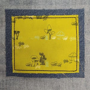May include: Close-up of a light blue denim fabric with a yellow patch. The patch features a black outline illustration of mushrooms and other plants. The patch is framed by a dark blue border.
