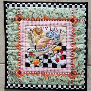 May include: A colorful quilted wall hanging with a black and white checkered border. The center features a whimsical illustration of a hat, flowers, and a sunflower with the text "Sunny Days".