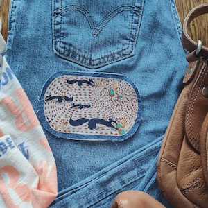 May include: Light blue denim jeans with a patch featuring a fox design. The patch is oval-shaped with a cream background and a pattern of foxes and foliage. A brown leather purse, a pair of brown flats, and a t-shirt are also visible.