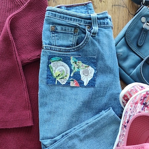May include: A pair of blue denim jeans with a patch pocket on the front. The pocket has a colorful floral and butterfly design.