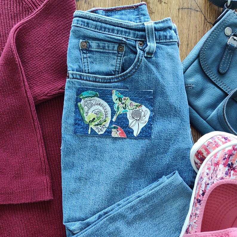 May include: A pair of blue denim jeans with a patch pocket on the front. The pocket has a colorful floral and butterfly design.