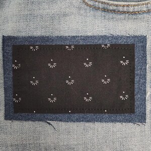 May include: Close-up of light blue denim jeans with a black patch. The patch is decorated with a white floral pattern. The jeans have a button closure and a pocket visible.