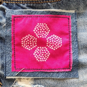 May include: A pink fabric patch with a white flower design sewn onto a pair of blue denim jeans.