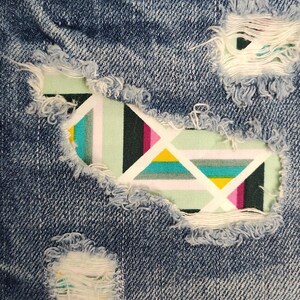May include: Blue denim jeans featuring multiple distressed areas with frayed edges. Behind the rips, a fabric patch is visible, displaying a geometric pattern with stripes in shades of mint green, black, yellow, white, and magenta.