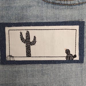 May include: Light blue denim jeans with a rectangular patch featuring black cactus illustrations. The patch is bordered by a dark blue stitched frame. The design includes a large cactus and two smaller cacti.