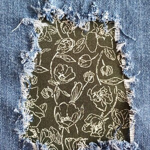 May include: Blue denim fabric featuring a rectangular distressed cutout with frayed edges. Beneath the opening, a dark olive green fabric patch is visible, decorated with a white line-art pattern of various blooming flowers and stems.