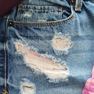 May include: Close-up of a pair of blue denim shorts with several distressed areas. The shorts have a front pocket and a frayed hem.