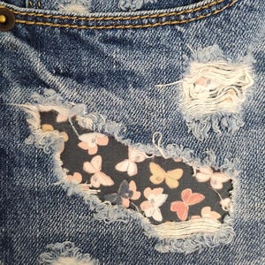 May include: Blue denim jeans featuring distressed areas with frayed edges. A dark fabric patch with a repeating pattern of small pink, white, and yellow butterflies is visible through a large horizontal rip on the front of the garment.