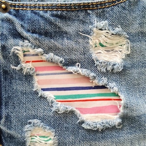 May include: Blue denim jeans featuring distressed areas with frayed edges. Beneath the rips, a colorful fabric patch is visible, displaying horizontal stripes in shades of pink, blue, green, and white. The denim has a classic wash and gold rivets.