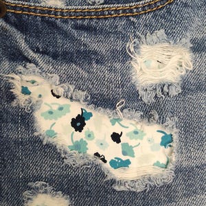 May include: Blue denim jeans featuring distressed areas with frayed edges. A white fabric patch with a floral pattern in shades of light blue, teal, and dark navy is visible underneath the largest distressed section of the denim fabric.
