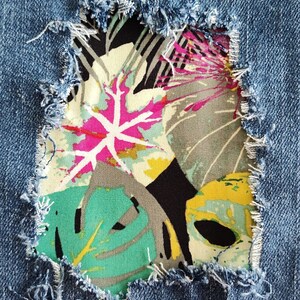 May include: A blue denim fabric featuring a distressed rectangular cutout with frayed edges. The opening reveals a colorful fabric patch underneath, decorated with a vibrant tropical leaf pattern in shades of pink, green, yellow, and black.