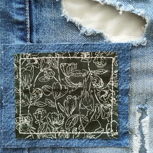 May include: A pair of blue denim jeans with a floral patch sewn over a hole. The patch is a dark green fabric with a white floral pattern.