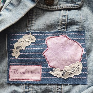 May include: A denim jacket with a patch that says "Floral Elements" and features a pink floral design and white lace accents.