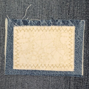 May include: A rectangular fabric patch with a cream-colored center featuring a subtle pattern, framed by a denim border. The edges are stitched with a zigzag pattern in a gold color, creating a decorative element. The patch is set against a denim background.