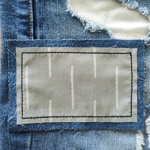 May include: A rectangular gray fabric patch with a white vertical dashed line pattern is stitched onto blue distressed denim jeans. The patch features a dark navy blue border of straight stitches securing it to the frayed denim fabric.