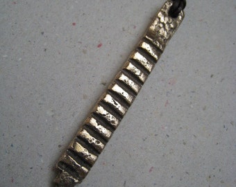 May include: A gold-toned pendant necklace with a long, rectangular pendant featuring a textured, grooved design. The pendant is attached to a black leather cord.