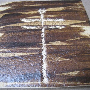 Pode incluir: Um azulejo de cerâmica quadrado com um esmalte marrom e branco. O azulejo tem um desenho de cruz branca no centro.