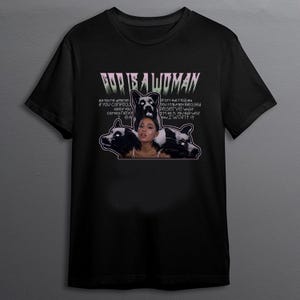 Op de afbeelding: Zwart t-shirt met een grafisch ontwerp. Het ontwerp toont de tekst "GOD IS A WOMAN" in een gestileerd lettertype, samen met een afbeelding van een vrouw en twee hondenillustraties. Er is ook extra tekst aanwezig.