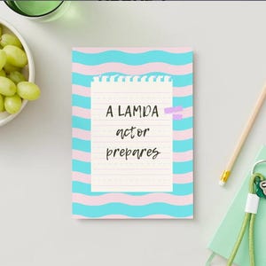 Puede incluir: Un bloc de notas con el texto "A LAMDA actor prepares" está pegado a una superficie blanca. El bloc de notas tiene un patrón ondulado rosa y azul. Un lápiz, un pequeño cuaderno verde y un cuenco de uvas verdes también están en la superficie.