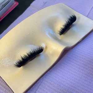 Pode incluir: Uma almofada de silicone bege em formato de rosto com dois conjuntos de cílios sintéticos pretos longos aplicados nas áreas dos olhos. A superfície da almofada é lisa e moldada para representar a ponte do nariz e as órbitas oculares.