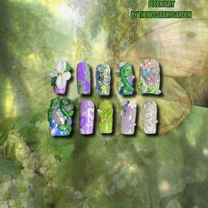 Puede incluir: Una colección de diez uñas artificiales con varios diseños. Las uñas presentan una gama de colores, incluyendo morado, verde y blanco, con adornos como flores, purpurina y mariposas. El texto "PIXIE FAIRY BY THEMOSSFAIRYGARDEN" es visible.