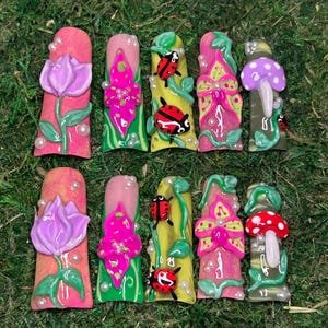 May include: A set of ten artificial nails with various designs. The nails feature vibrant colors and 3D embellishments, including flowers, ladybugs, and mushrooms. Each nail is uniquely decorated with intricate details and pearl accents, set against a green background.