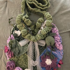 Può includere: Borsa all'uncinetto fatta a mano nei toni del verde, viola e blu, ornata con motivi floreali e farfalle. La borsa presenta una lunga tracolla ad anello ed è legata con un nastro di pizzo bianco. Un accessorio unico e artigianale.