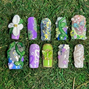 Puede incluir: Diez uñas artificiales con diversos diseños. Las uñas muestran colores como morado, verde y blanco, adornadas con purpurina, acentos florales y detalles de mariposas. Están dispuestas sobre musgo verde. Cada uña tiene un diseño único.
