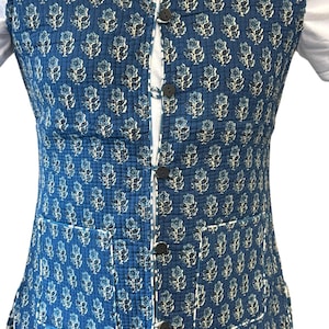 May include: A blue quilted waistcoat featuring a floral design in white and light blue. It has a mandarin collar, a button-up front, and two front pockets. The waistcoat is worn over a white shirt.
