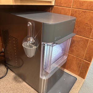 Op de afbeelding: Een roestvrijstalen ijsmachine met een doorzichtige ijsbak en een grijze bovenkant. Een doorzichtige plastic schep hangt aan de zijkant. De ijsmachine staat op een aanrecht naast een betegelde muur.