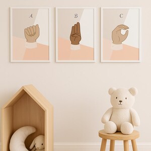 Op de afbeelding: Drie ingelijste prints met handgebaren die de letters A, B en C in gebarentaal spellen. Een houten huisvormige plank bevat een maansikkelkussen en een knuffelkonijn. Een teddybeer zit op een kleine houten kruk.
