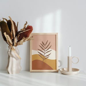 Op de afbeelding: Een stilleven met een beige vaas met gedroogde bloemen, een ingelijste abstracte kunstdruk en een kandelaar met een brandende witte kaars. De kunstdruk heeft een bruin bladerenpatroon.