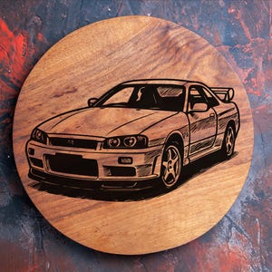 Puede incluir: Una placa redonda de madera con una ilustración grabada de un coche deportivo. El coche se muestra de perfil, destacando su diseño y características. La madera tiene una veta natural.