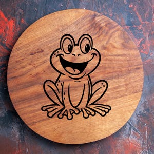 May include: A round wooden cutting board with a smiling frog illustration. The frog is black and has large eyes and a wide grin. The board is made of wood and has a natural wood grain pattern.