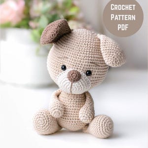 Peut inclure: Un jouet pour chien crocheté beige et marron. Le chien a une tête ronde, des oreilles tombantes et un petit nez marron. Le texte "CROCHET PATTERN PDF" est dans un cercle en haut à droite. Le jouet est posé sur une surface blanche.