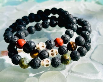 Puede incluir: Tres pulseras apiladas hechas con cuentas de lava negra, cuentas rojas, cuentas de ojo de tigre, cuentas verdes y cuentas de calavera blancas.