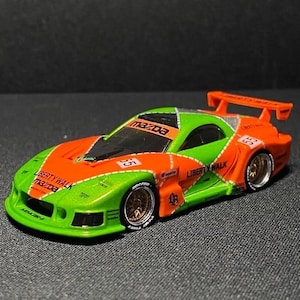 Puede incluir: Un coche de juguete Mazda RX-7 verde y naranja con el texto "Liberty Walk" y "Mazda" impreso. El coche de juguete tiene llantas doradas y un alerón trasero. El coche está sobre una superficie gris oscuro.