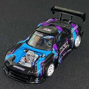 Op de afbeelding: Een zwart modelauto met een blauw en paars verfspatontwerp. De motorkap staat open en toont een gedetailleerde motorruimte. Het model is voorzien van een grote achtervleugel, witte wielen en HKS- en HIPERMAX-branding op de carrosserie.