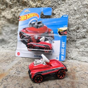 Könnte beinhalten: Rotes Hot Wheels Pedal Drive Ride-On Spielzeugauto mit schwarzen Rädern und Flammen an der Seite. Das Auto hat ein weißes Lenkrad und einen schwarzen Motor. Die Verpackung zeigt das Auto auf blauem Hintergrund mit dem Text "Hot Wheels Pedal Drive" und "HW Ride-Ons".