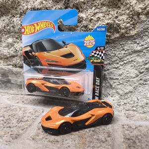 Könnte beinhalten: Orange-schwarzes Hot Wheels McLaren Spielzeugauto. Das Auto wird in der Originalverpackung präsentiert, mit dem Jahr 2025 auf der Verpackung. Ein zweites Auto wird außerhalb der Verpackung gezeigt.