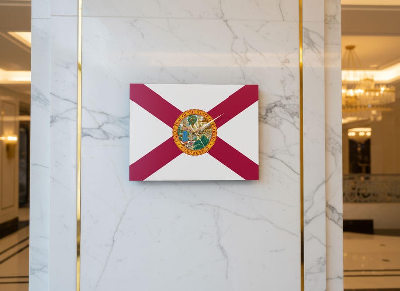K&ouml;nnte beinhalten: Eine rechteckige Wanduhr mit dem Design der Flagge von Florida. Das Zifferblatt ist wei&szlig; mit roten diagonalen Streifen und einem kreisf&ouml;rmigen Emblem in der Mitte. Goldene Zeiger zeigen die Uhrzeit an.