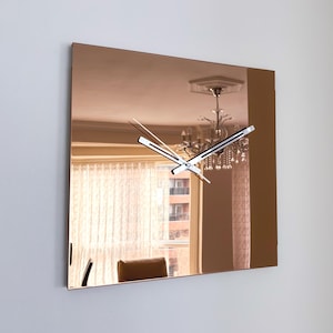 May include: Square copper-toned wall clock with a mirrored surface. The clock features silver hands and a minimalist design. The reflection shows a chandelier and a window with curtains. The clock is mounted on a white wall.