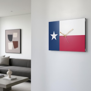 Könnte beinhalten: Quadratische Wanduhr mit dem Design der Texas-Staatsflagge. Das Zifferblatt ist in blaue, weiße und rote Abschnitte unterteilt, mit einem weißen Stern auf der blauen Seite. Goldfarbene Zeiger zeigen die Uhrzeit an. Die Uhr ist an einer weißen Wand befestigt.