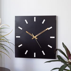 May include: Square black wall clock with gold-colored hands and white rectangular markers. The clock is mounted on a white wall, with green plants in the foreground.
